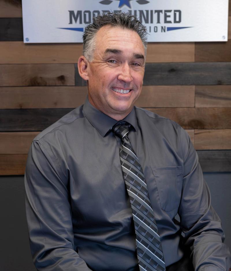 Smiling team member in a gray shirt and tie seated in front of the company sign