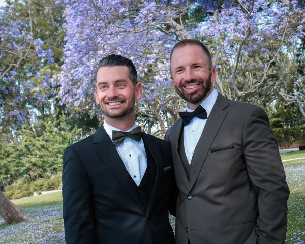 Grooms laughing under a canopy of jacaranda blooms