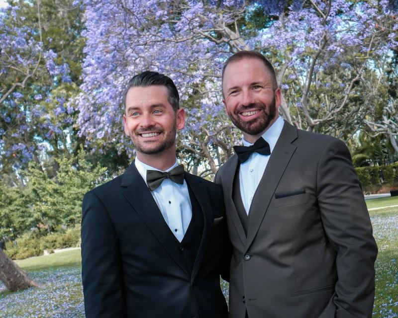Grooms laughing under a canopy of jacaranda blooms
