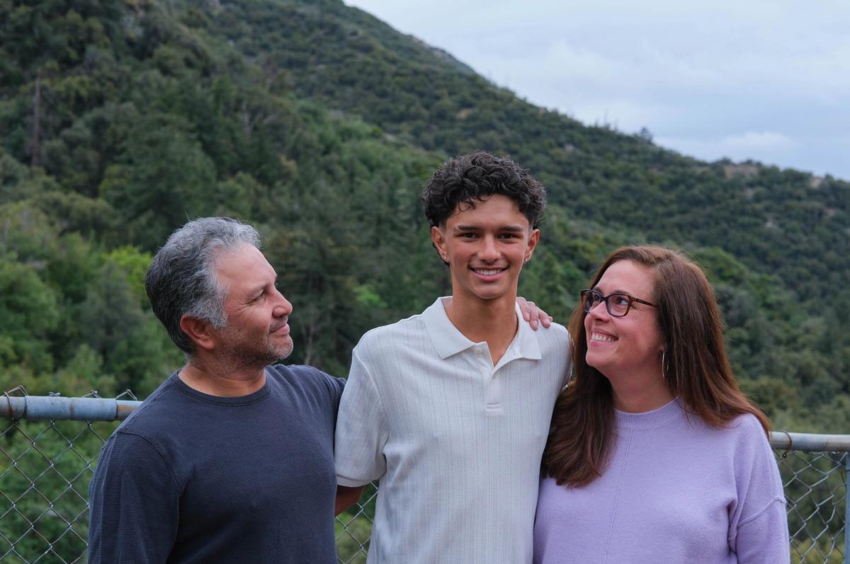 Teen son standing between his mom and dad with green mountains behind them