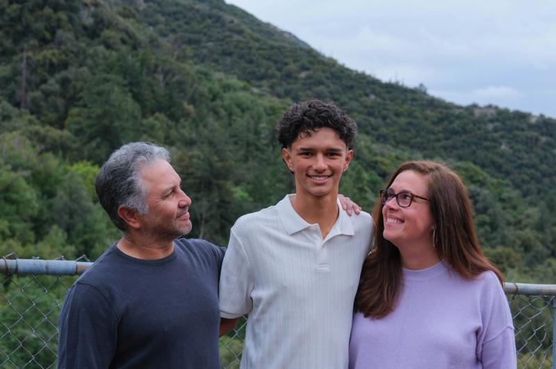 Teen son standing between his mom and dad with green mountains behind them