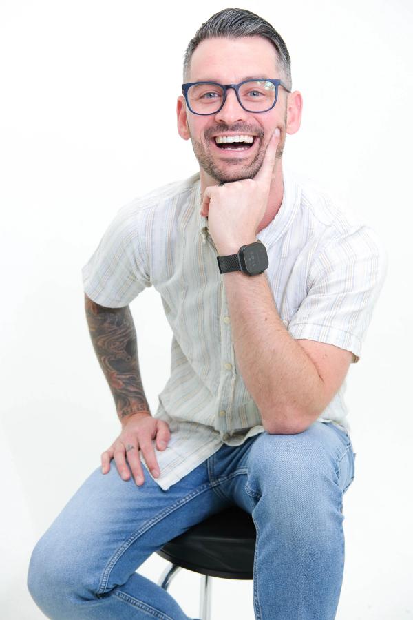 Man in glasses laughing with his hand on his chin, seated on a studio stool