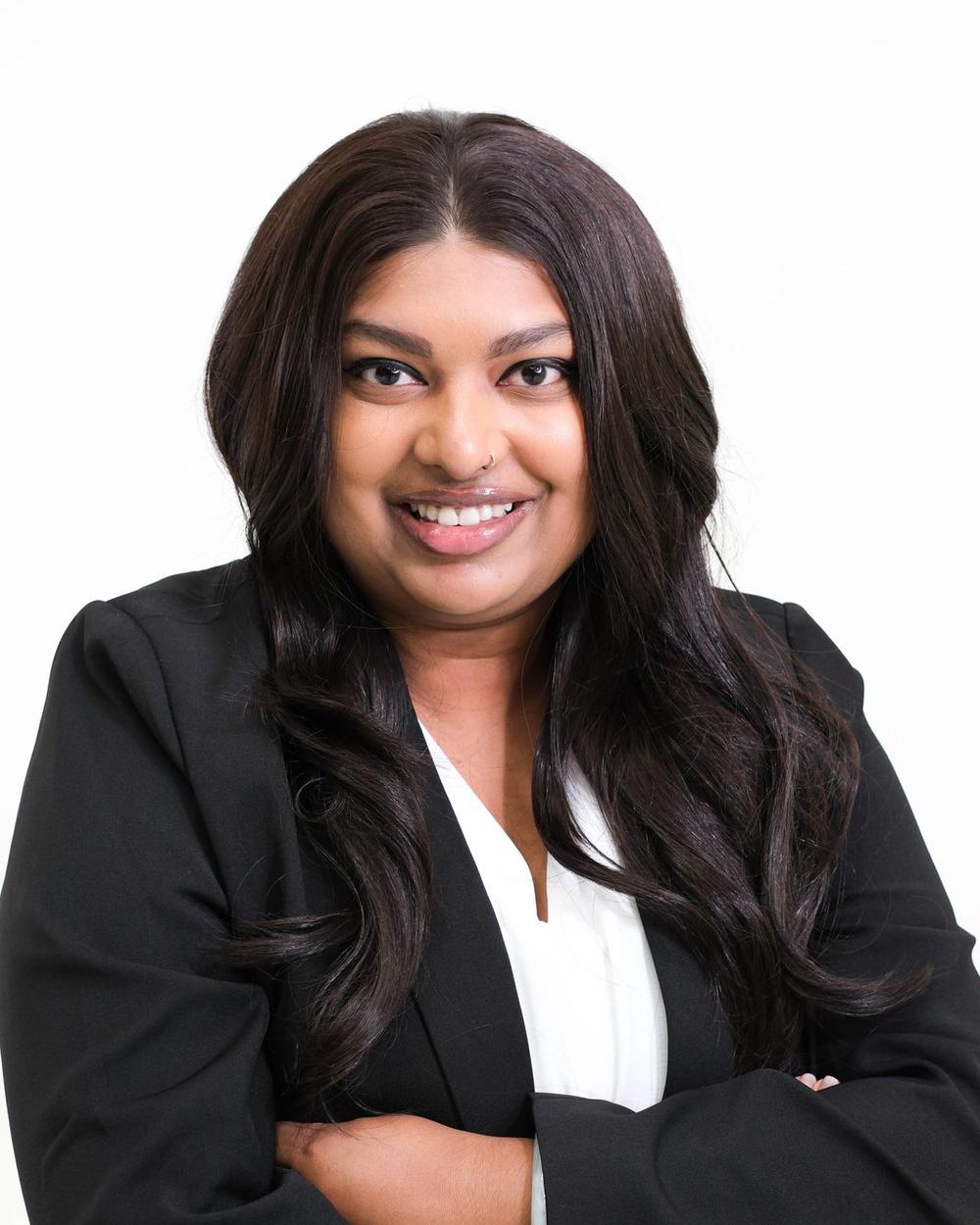 Professional woman in a black blazer and white blouse, arms crossed, smiling on a clean studio backdrop