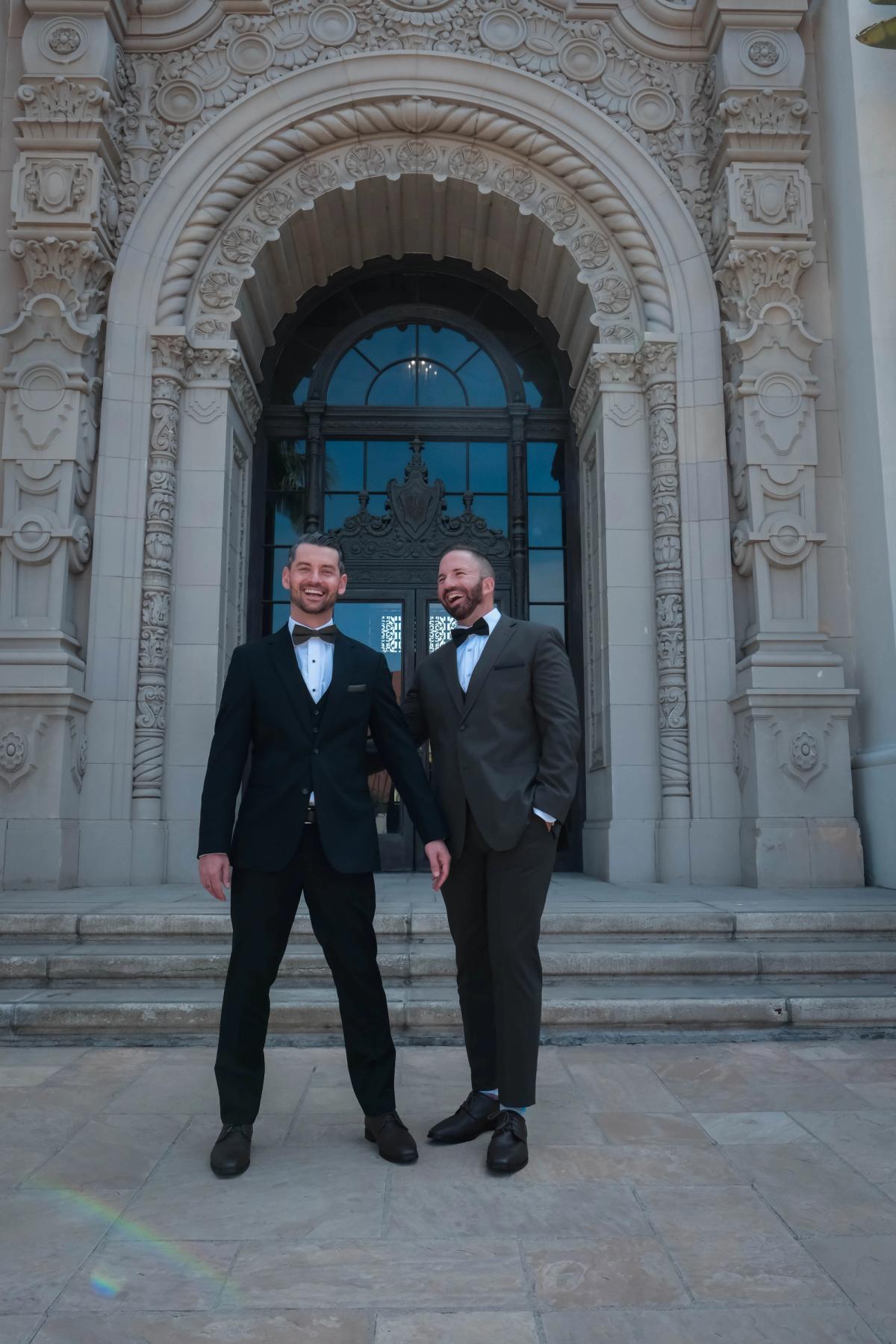 Two grooms in black tuxedos standing at the arched entrance of a historic city hall