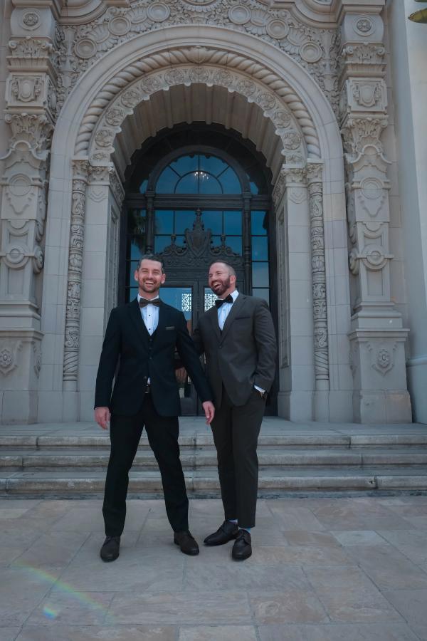 Two grooms in black tuxedos standing at the arched entrance of a historic city hall
