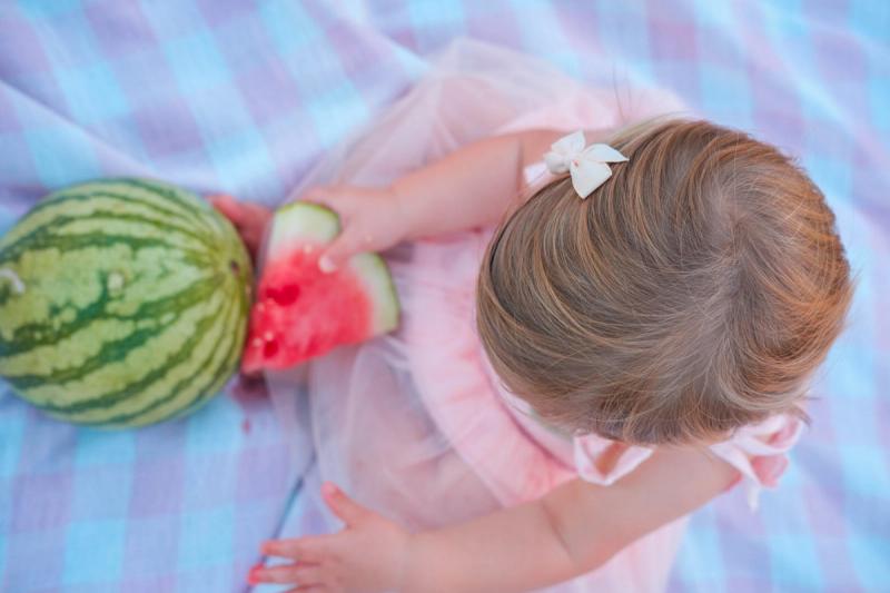 Overhead view of a toddler with a watermelon slice on a gingham blanket