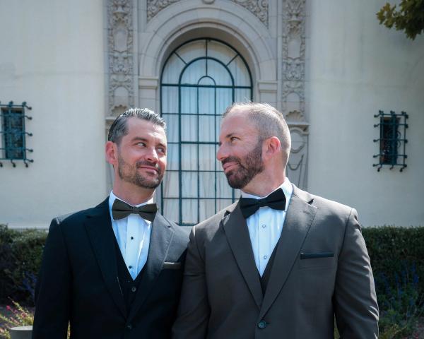 Grooms in tuxedos smiling at each other in front of an arched city hall window