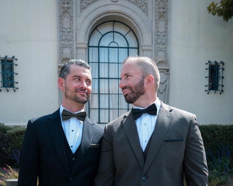 Grooms in tuxedos smiling at each other in front of an arched city hall window