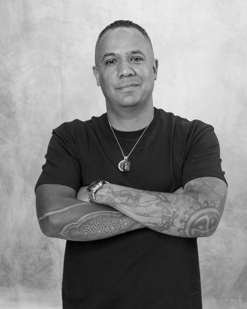 Man with tattooed arms crossed against a mottled gray backdrop