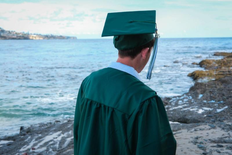 Graduate in cap and gown seen from behind, looking out at the ocean