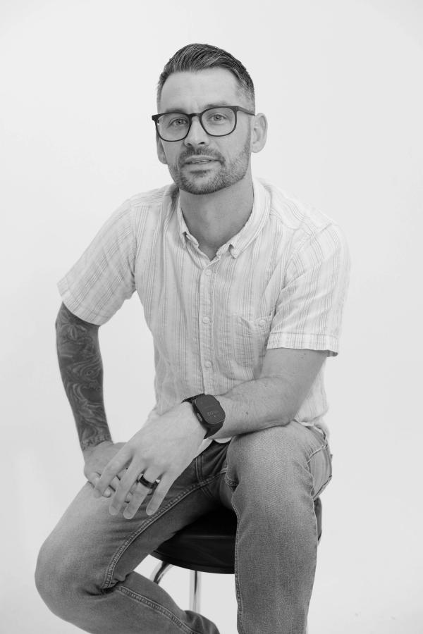 Black and white portrait of a man in glasses seated on a studio stool