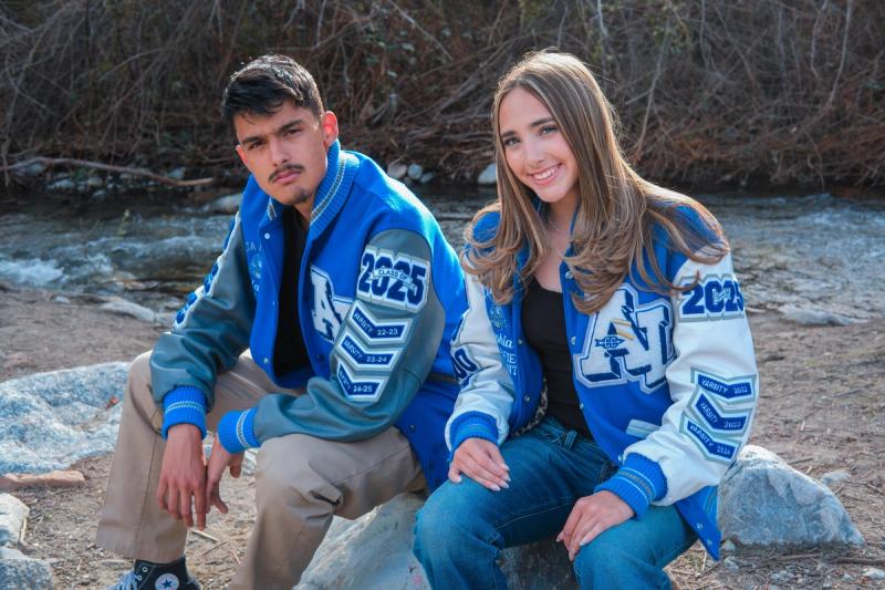 Senior siblings in letterman jackets sitting on a boulder by the water