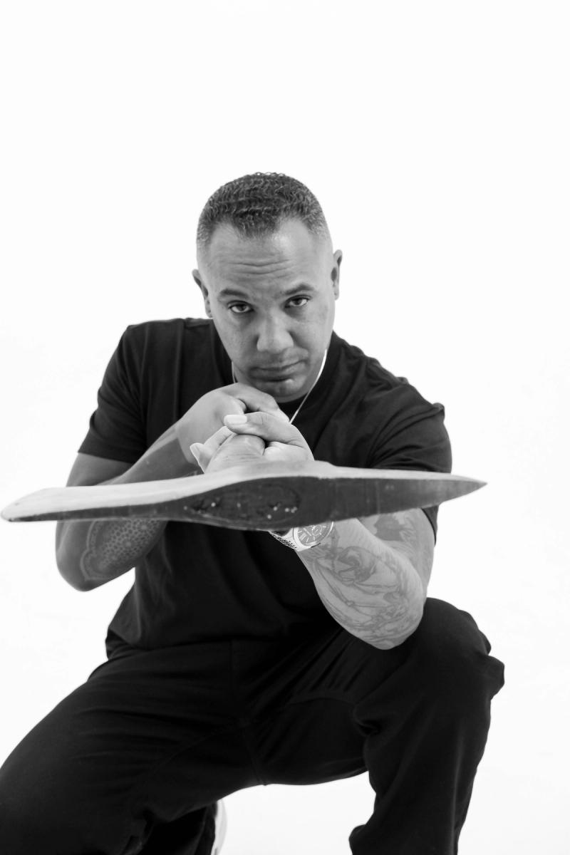 Black and white portrait of a man crouching and balancing a skateboard deck in front of him
