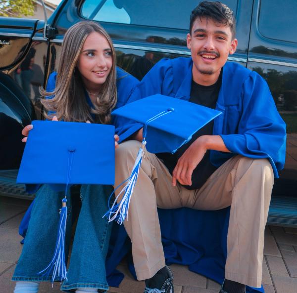 Two graduates in blue gowns sitting against a vintage black car holding their caps