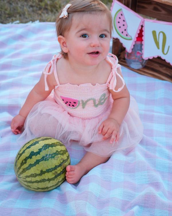 One-year-old girl in a pink tutu smiling beside a watermelon on a gingham blanket