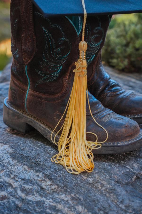 Detail of a graduation cap and gold tassel resting on cowboy boots