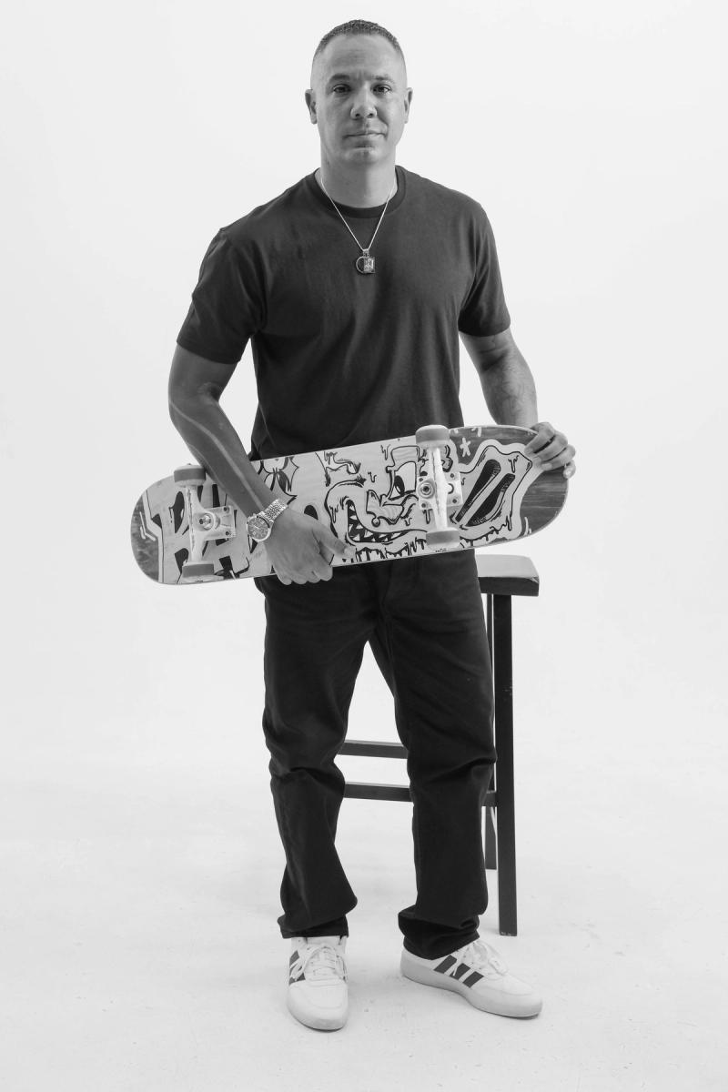 Man seated on a stool holding a graffiti skateboard deck across his lap