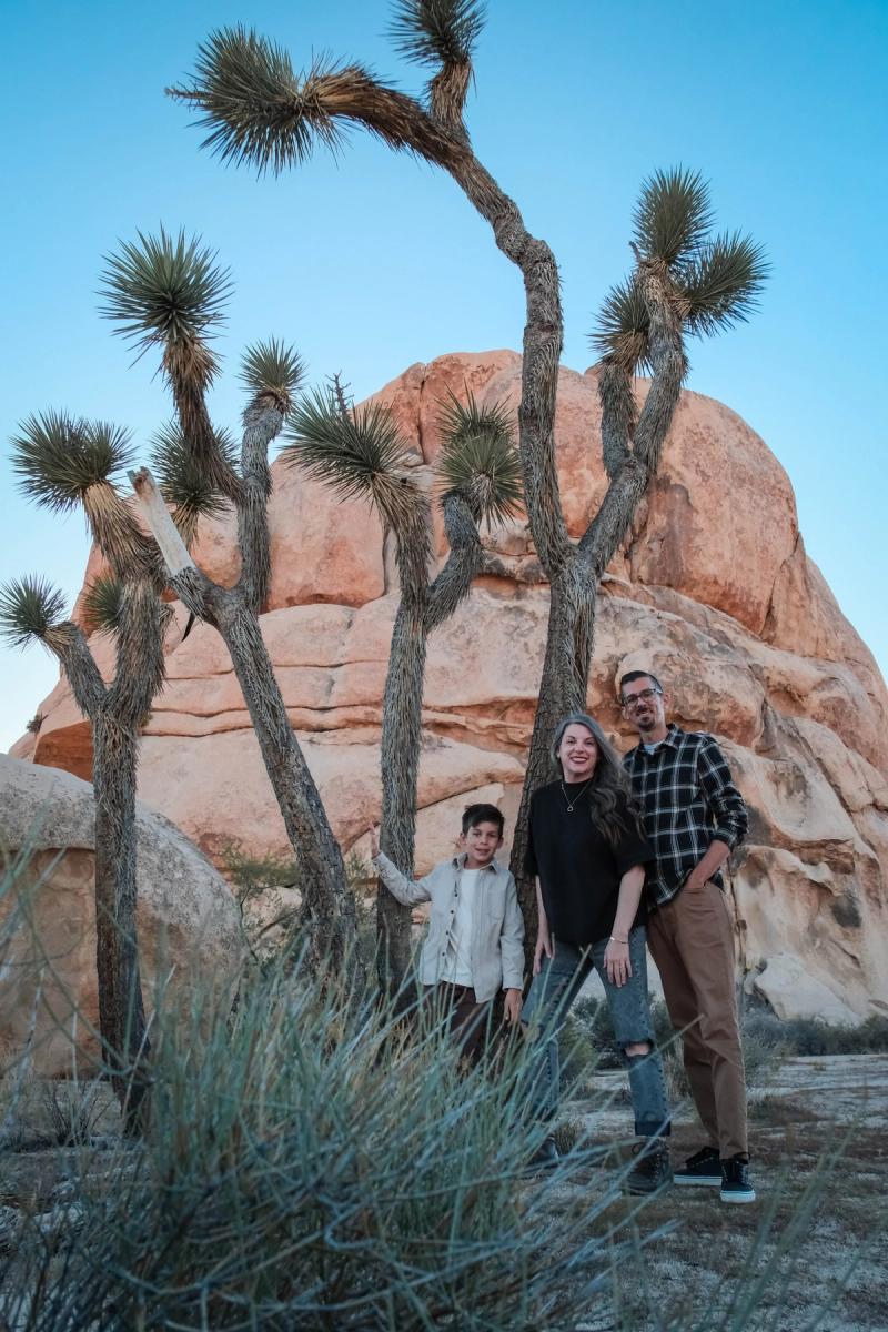 Family of three standing beneath tall Joshua trees and a red rock formation