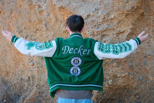 Senior facing a sandstone cliff with arms spread wide in his letterman jacket