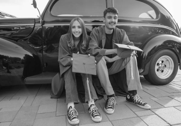 Two graduates in caps and gowns posed beside a classic black car