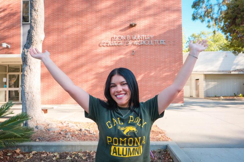 Graduate with arms raised in celebration in front of a brick campus building