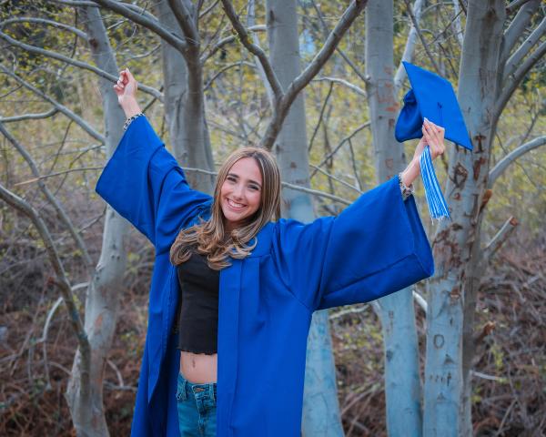 Graduate in a blue gown raising her arms and her cap among the trees
