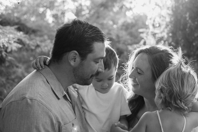 Black and white portrait of parents cuddling their son and daughter among the trees