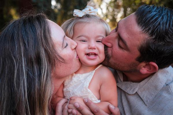 Mom and dad kissing their toddler daughter’s cheeks as she grins