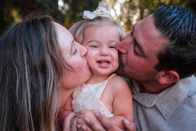Mom and dad kissing their toddler daughter’s cheeks as she grins