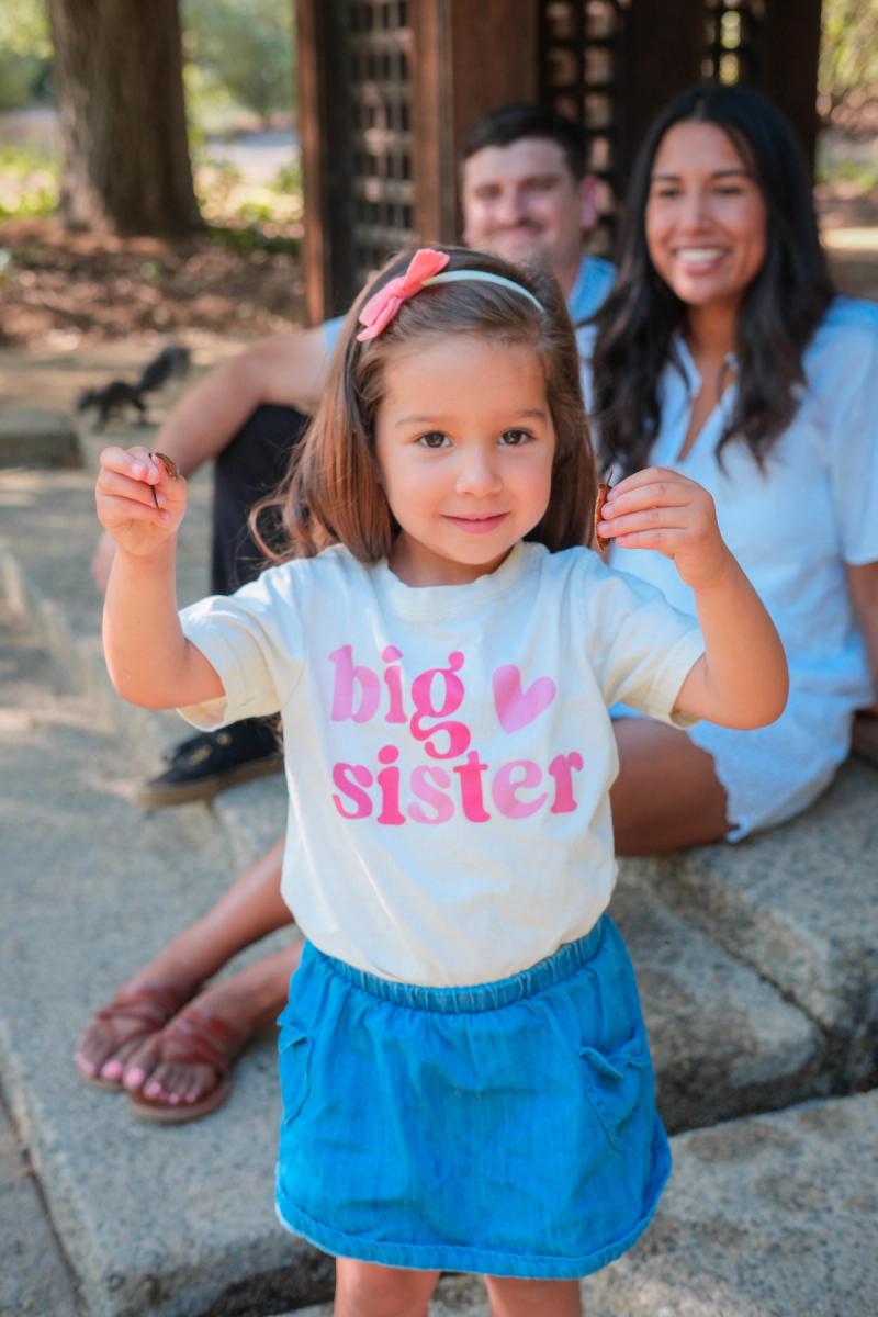 Little girl in a “big sister” shirt with her parents behind her