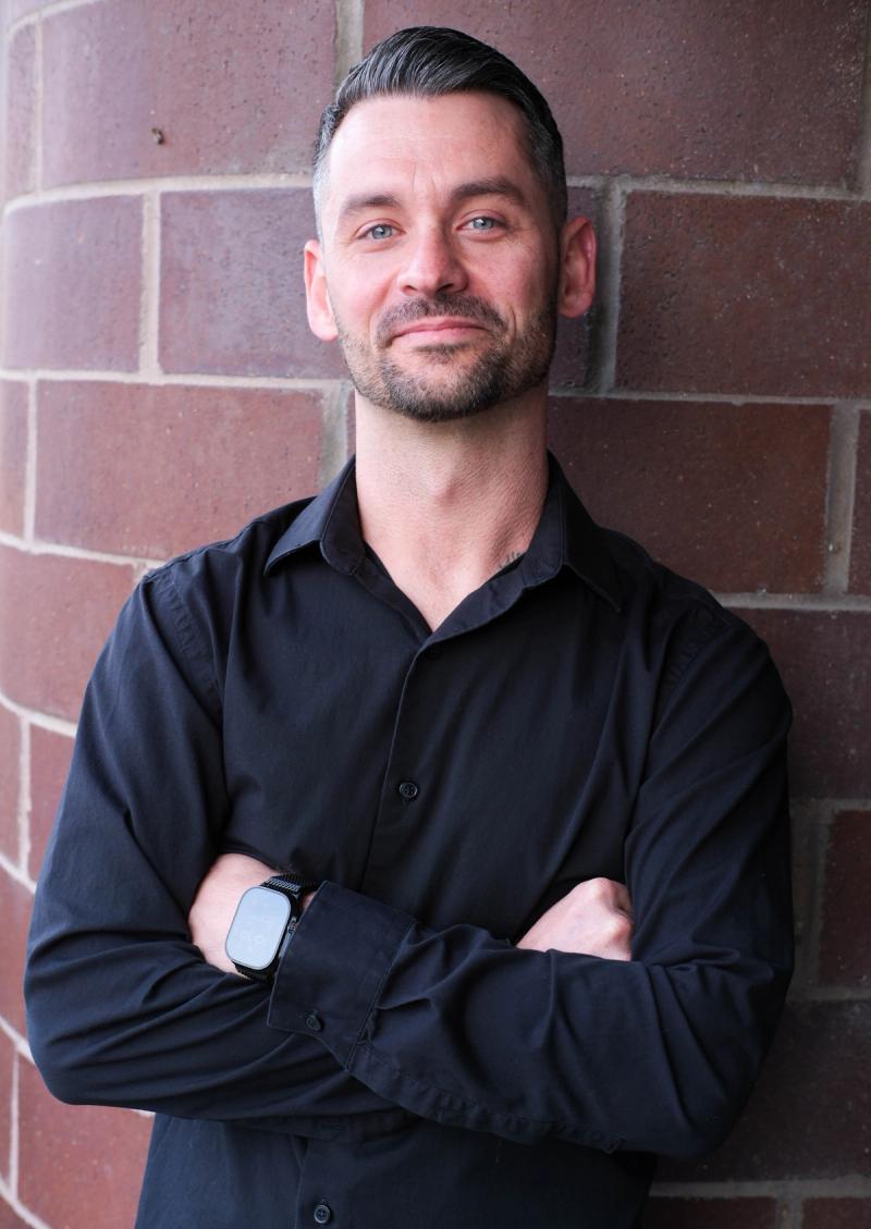 Man in a navy shirt with arms crossed, leaning against a red brick wall