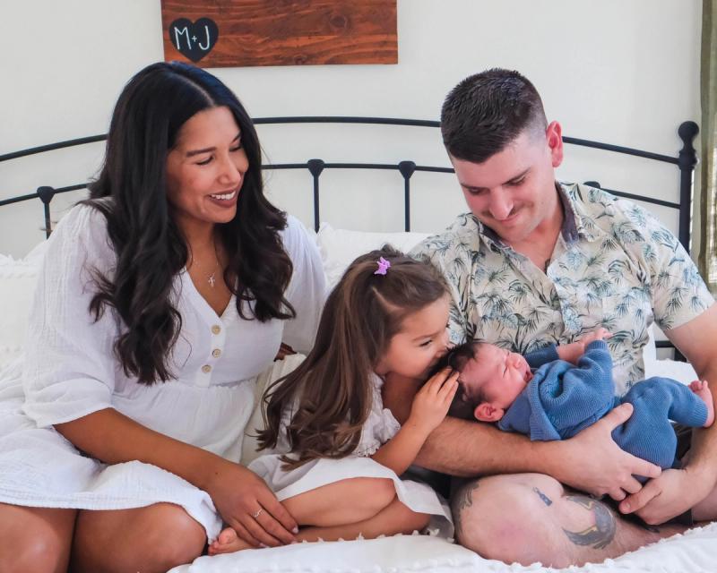 Parents and their daughter cuddling a newborn on a white bed