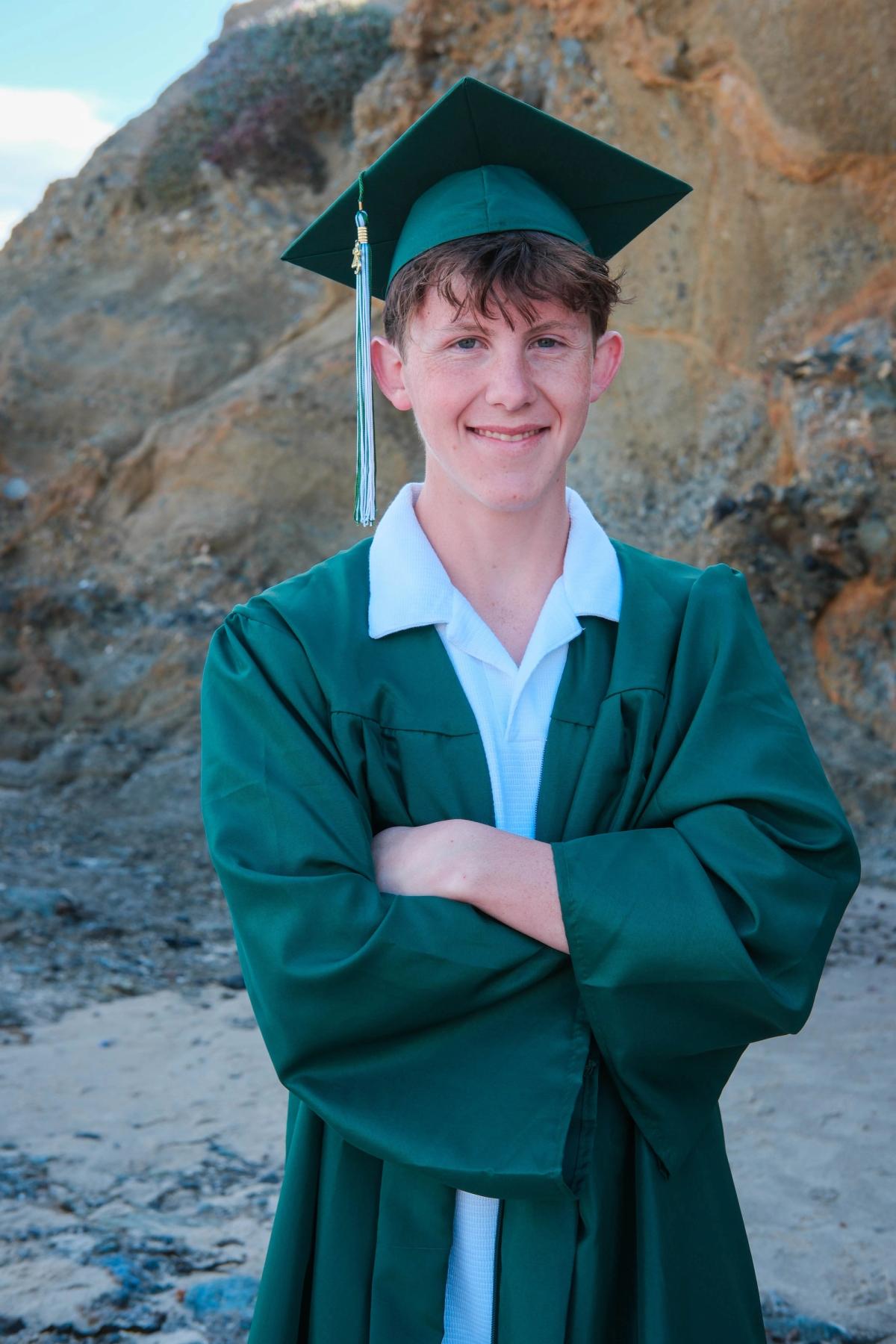 Smiling graduate in a green cap and gown with arms crossed in front of coastal rocks