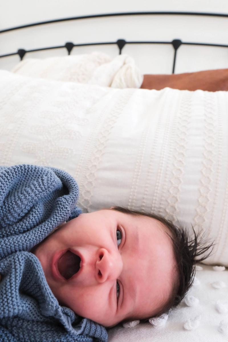 Newborn yawning in a blue knit sweater on a white bed