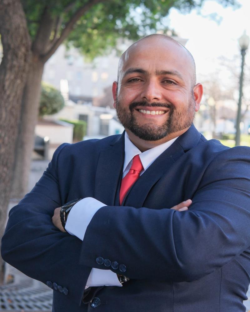 Smiling candidate in a navy suit and red tie with arms crossed outdoors