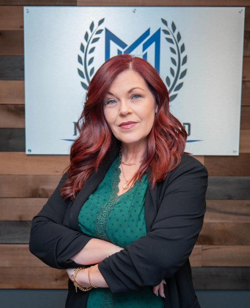 Team member in a green blouse and black blazer with arms crossed in front of the Moore United logo