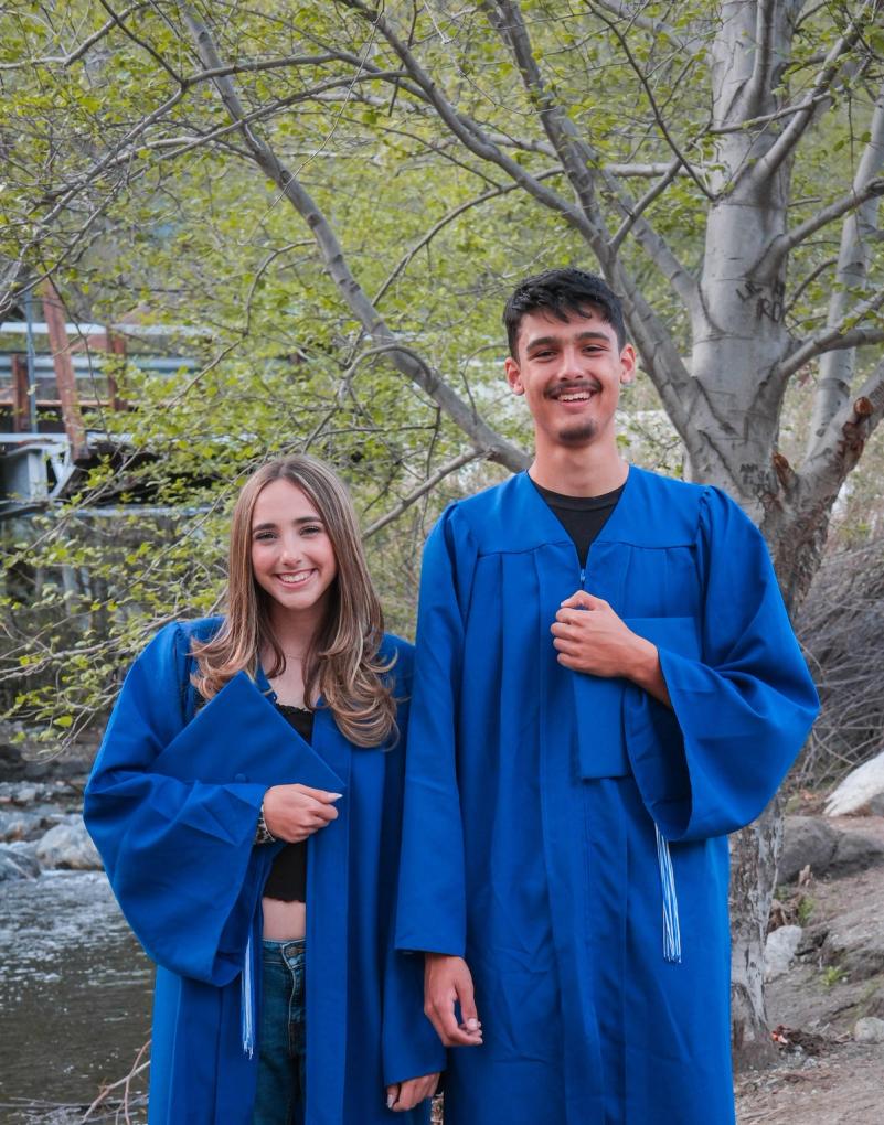 Brother and sister graduates in blue caps and gowns by a creek