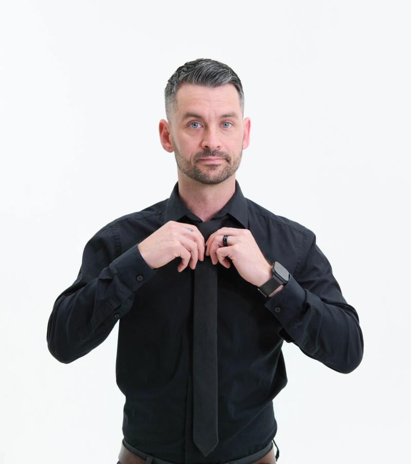 Man in a black shirt adjusting his tie, looking at the camera