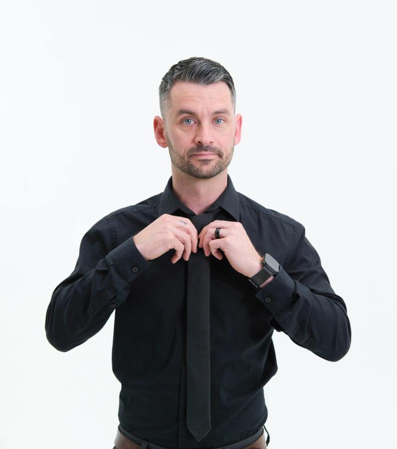 Man in a black shirt adjusting his tie, looking at the camera