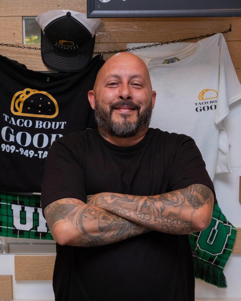 Owner with arms crossed beside Taco Bout Good shirts and hats