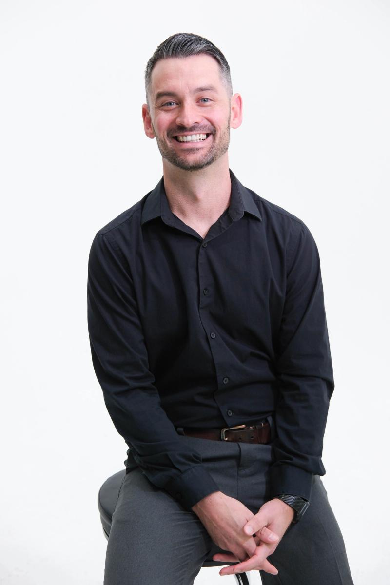 Smiling man in a black shirt seated on a stool with hands clasped