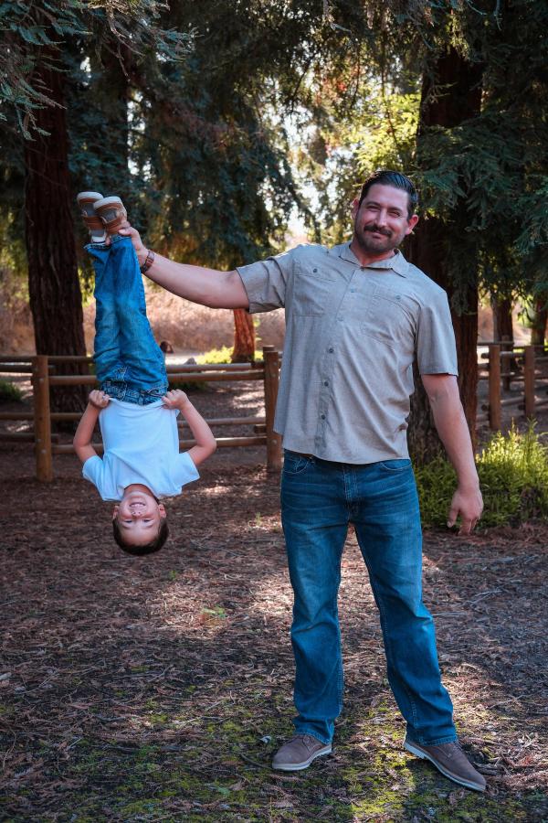 Dad holding his laughing son upside down by the ankles under the trees