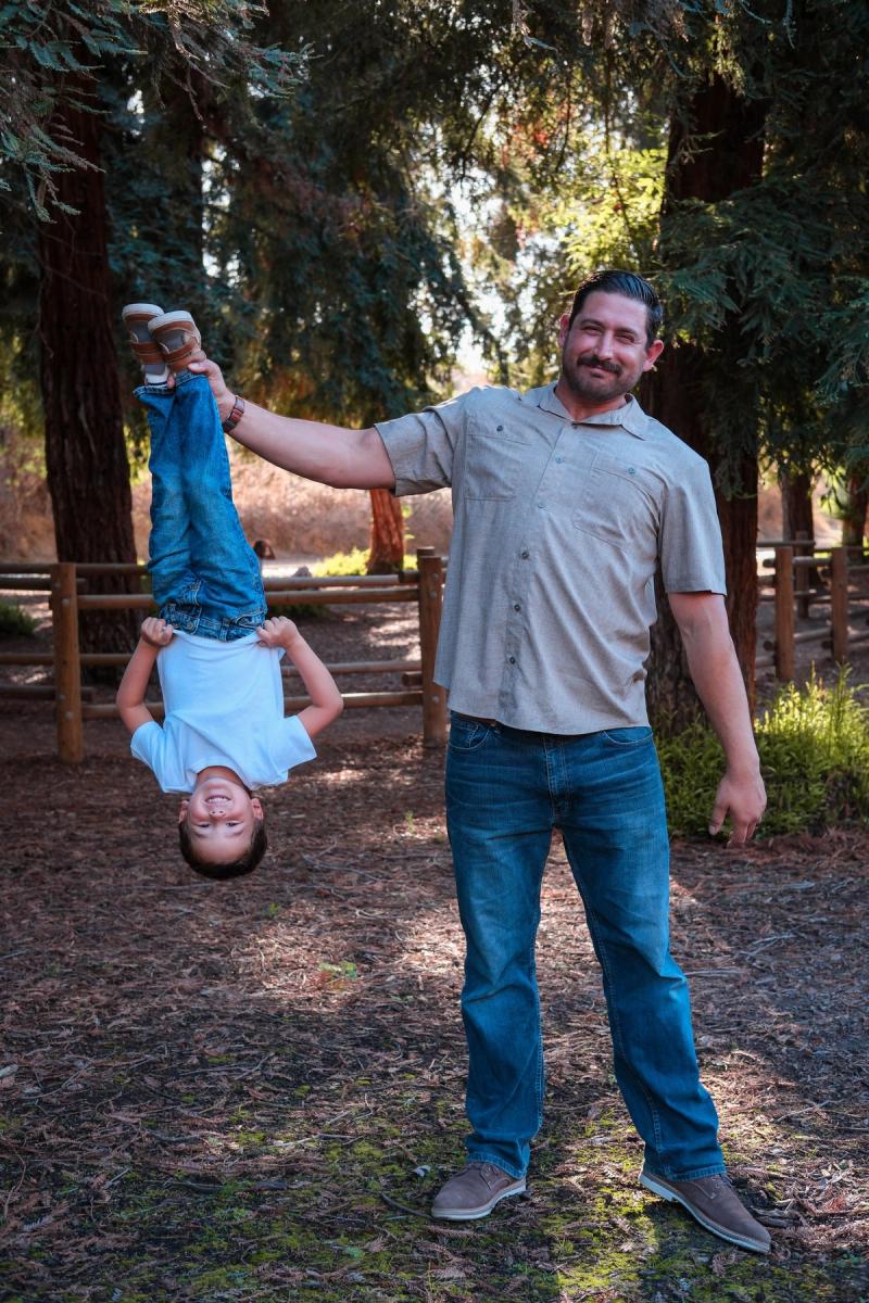 Dad holding his laughing son upside down by the ankles under the trees