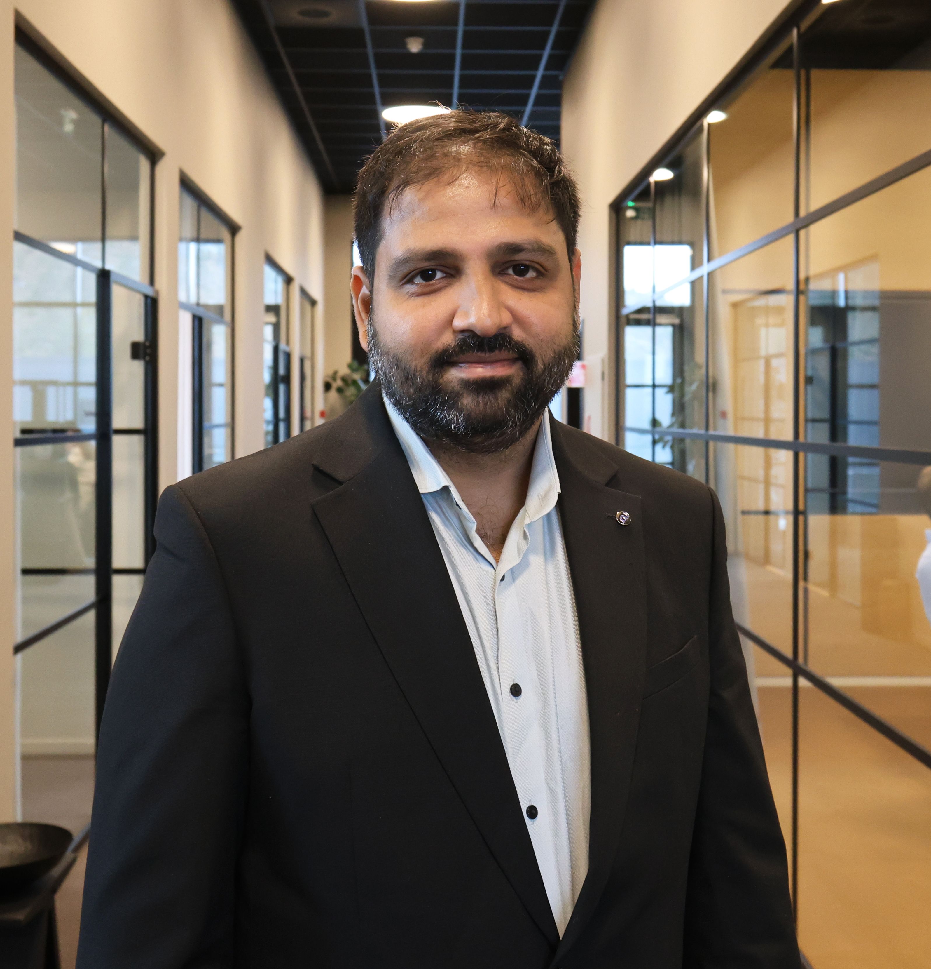 A bearded man in a black suit jacket and light blue shirt smiles in an office hallway.