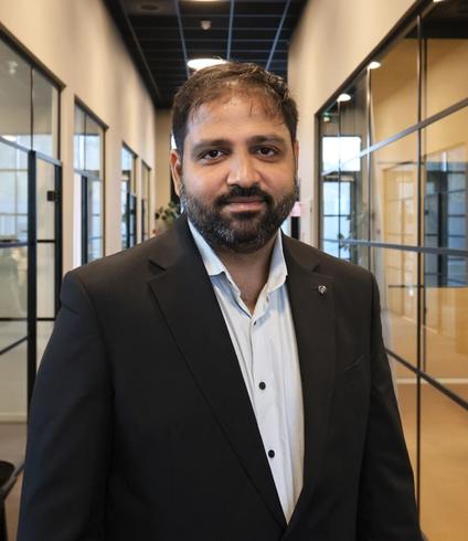 A bearded man in a black suit jacket and light blue shirt smiles in an office hallway.