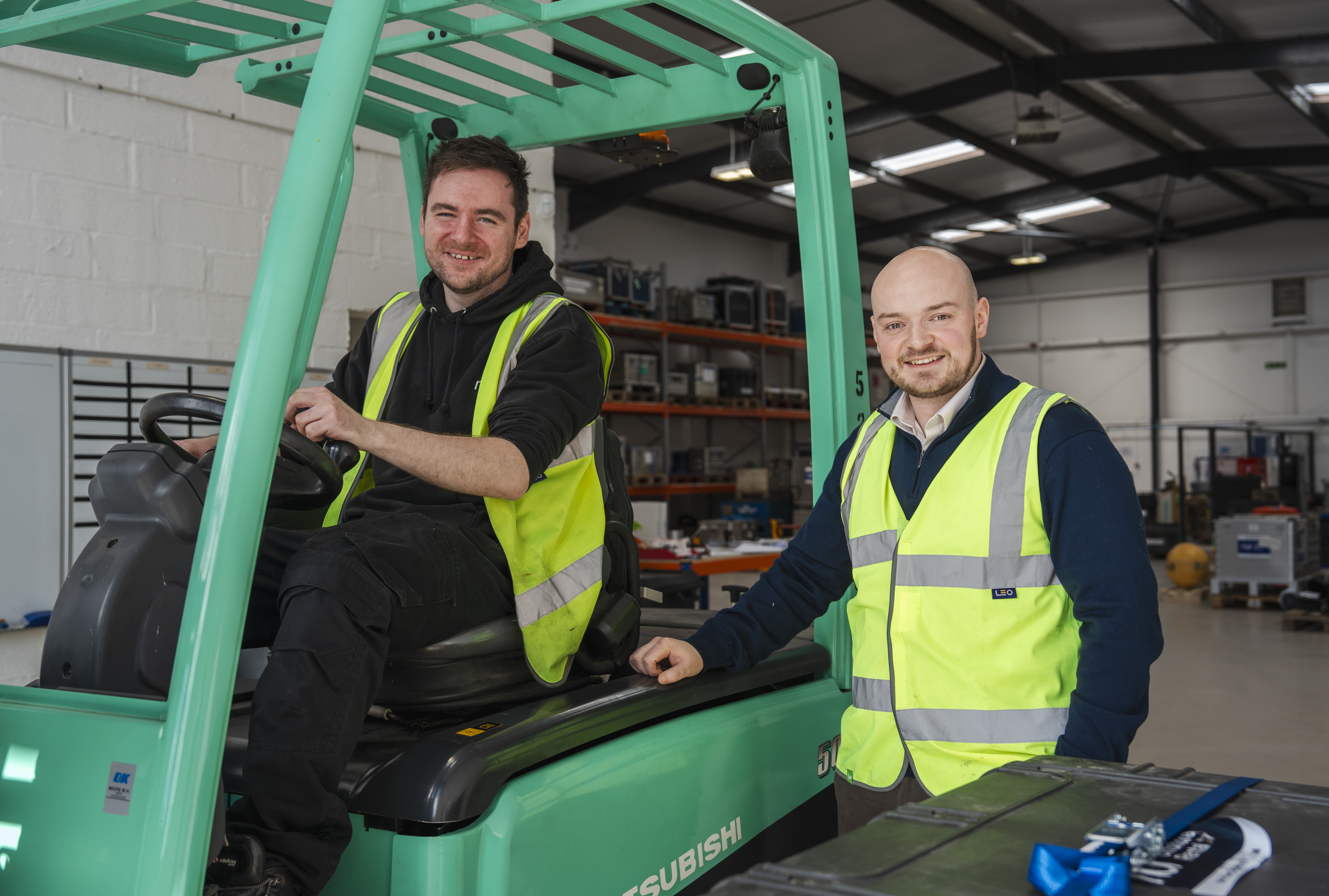 Two smiling men in hi-vis vests in a warehouse; one sits in a green forklift, the other stands beside it.