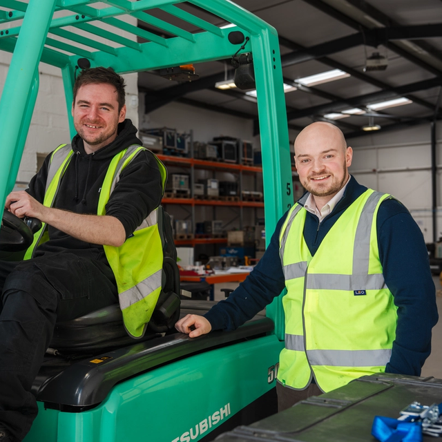 Two smiling men in hi-vis vests in a warehouse; one sits in a green forklift, the other stands beside it.