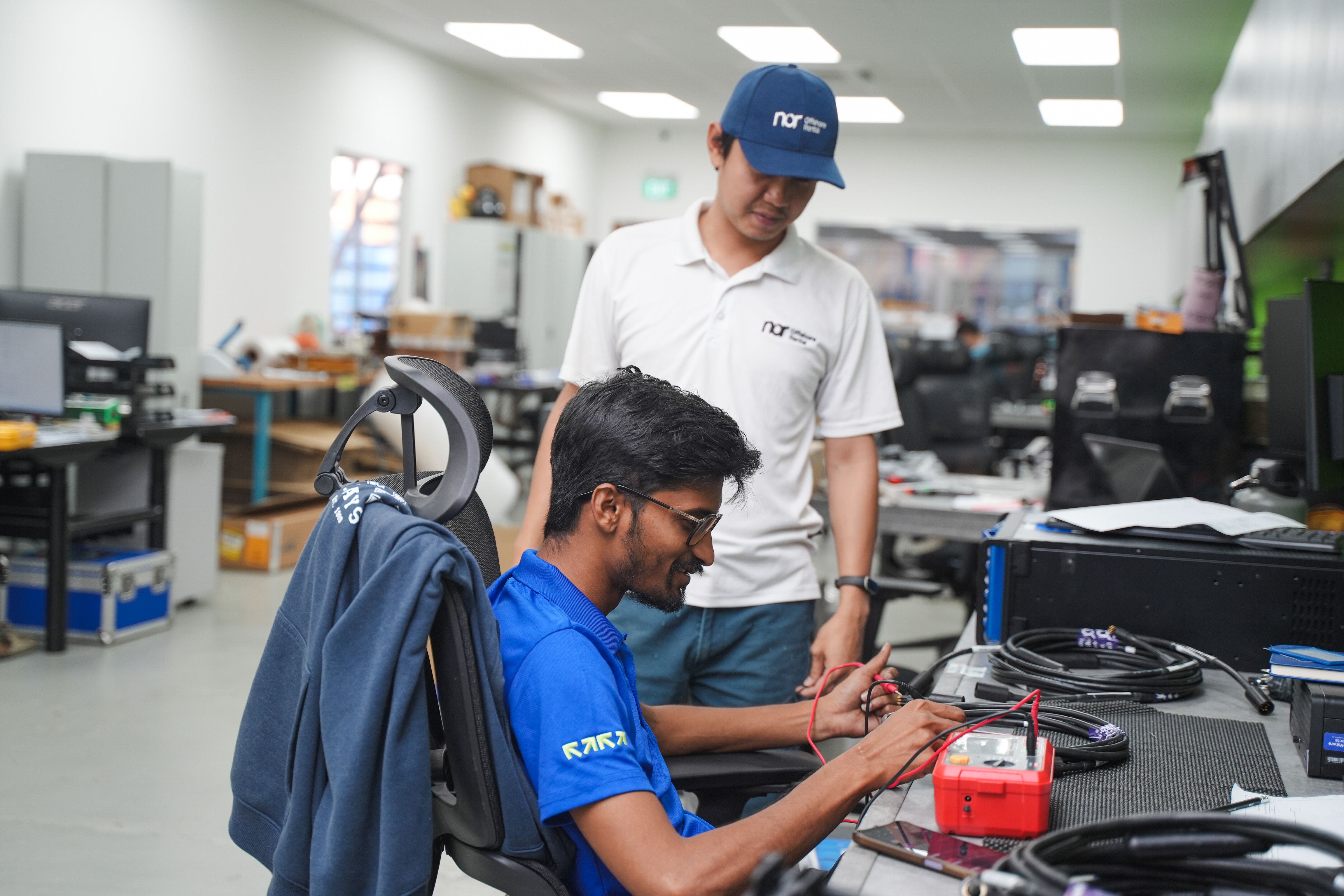 A man in a blue shirt tests cables with a multimeter, while another man in a white shirt and cap stands behind him, observing.