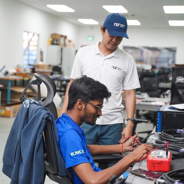 A man in a blue shirt tests cables with a multimeter, while another man in a white shirt and cap stands behind him, observing.