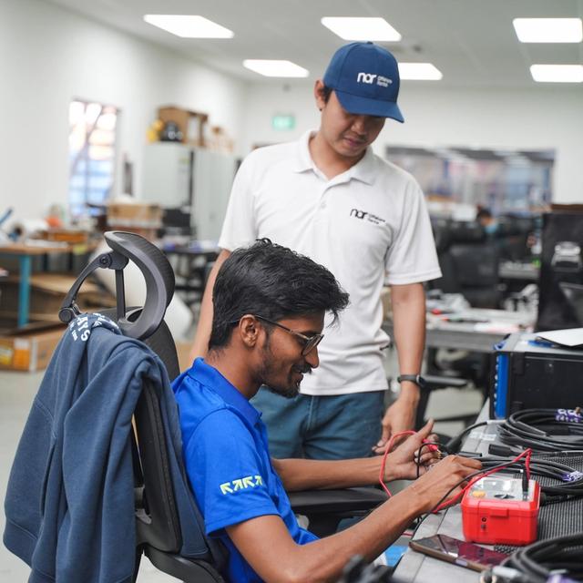 Two men in a lab, one seated and testing cables with a multimeter, the other observing.